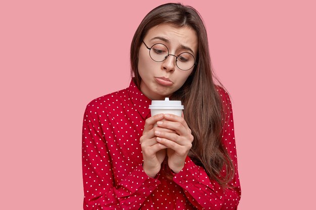 Mujer solitaria y abatida frunce el labio inferior, sostiene una taza desechable de bebida caliente, se siente triste, deprimida