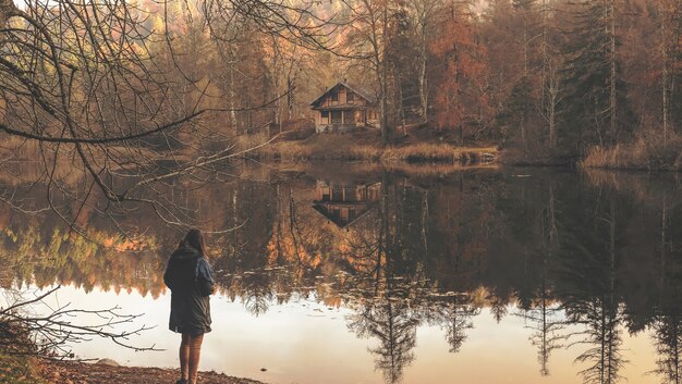 Mujer solitaria de pie cerca del lago con el reflejo de la cabaña de madera aislada visible