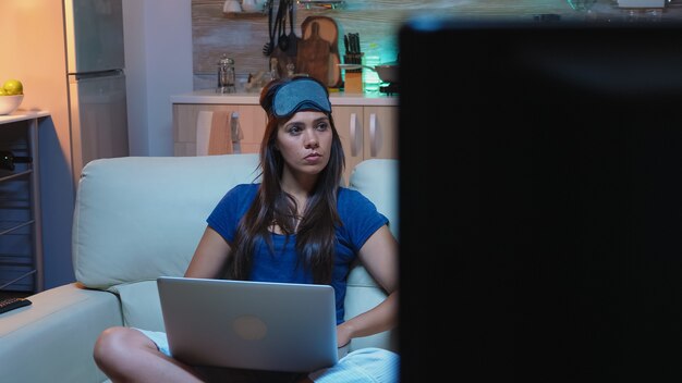 Mujer solitaria en pijama y máscara para dormir en la frente sentada en el sofá buscando información en la computadora portátil. Señora centrada escribiendo un correo electrónico utilizando tecnología de internet a altas horas de la noche en la sala de estar