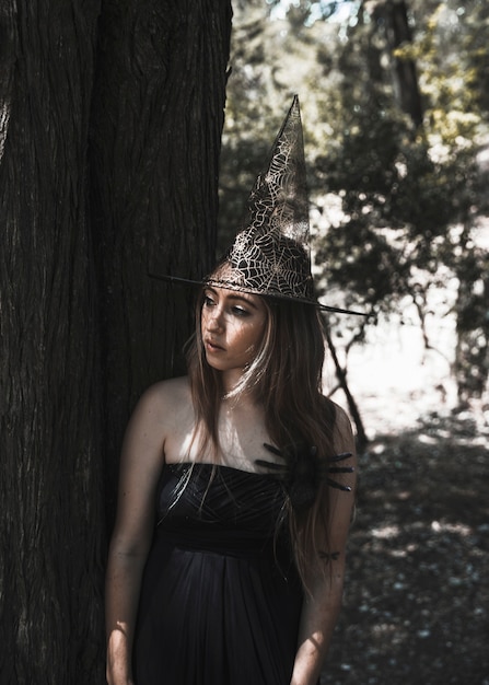 Mujer con sombrero de bruja de pie al lado del árbol y mirando a otro lado
