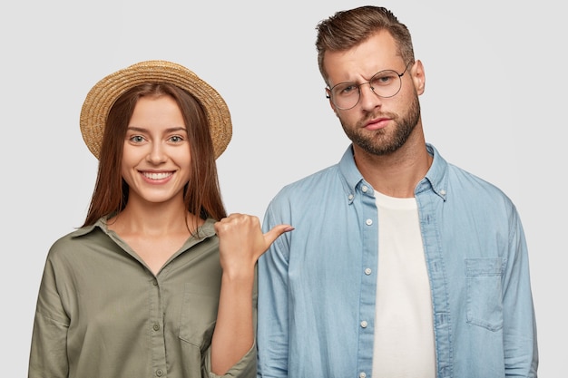 La mujer con sombrero de paja tiene una amplia sonrisa brillante, señala a su novio que tiene una expresión insatisfecha, pasan tiempo libre juntos, posan contra la pared