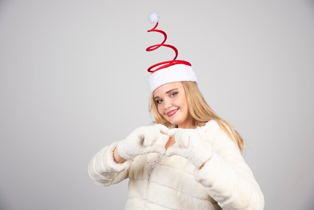 Mujer con sombrero de Santa haciendo signo de corazón en la pared gris.