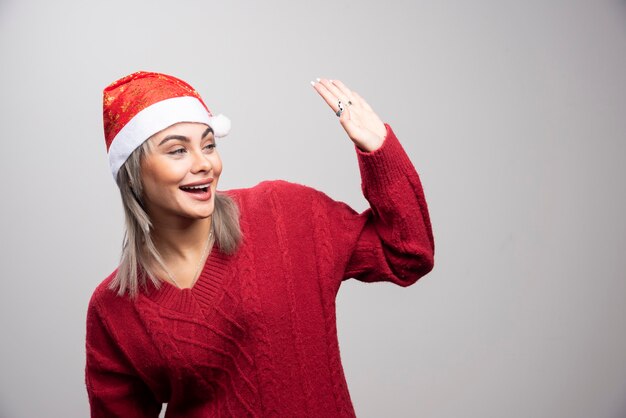 Mujer con sombrero de Santa saludando a alguien sobre fondo gris.