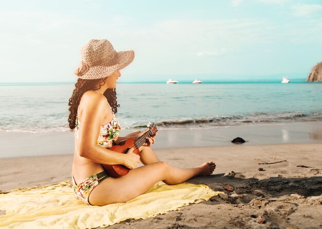 Mujer con sombrero tocando el ukelele en la playa de arena