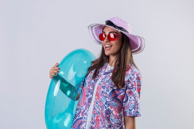 Mujer con sombrero de verano y gafas de sol rojas sosteniendo un anillo inflable mirando alegremente positivo y feliz sonriendo alegremente de pie en blanco aislado