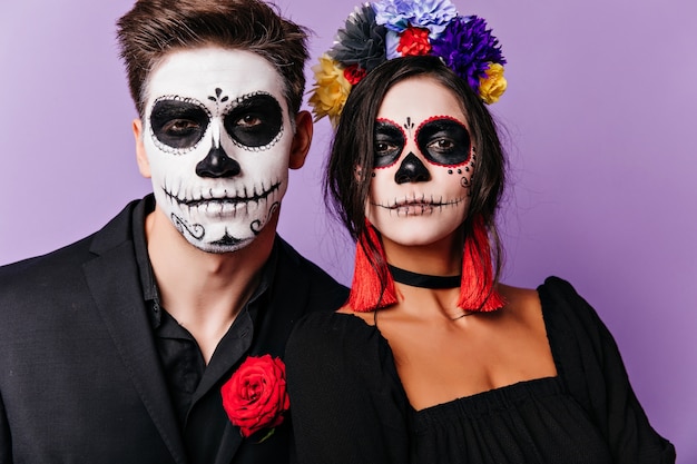 Mujer soñadora en corona de flores posando en halloween con novio. Chicos caucásicos en trajes de zombies de pie sobre fondo morado.