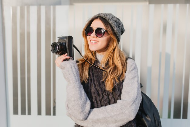 Mujer soñadora en gafas de sol con cámara en mano mirando hacia arriba con una sonrisa encantadora. Retrato al aire libre de fotógrafa inspirada con sombrero y suéter de punto suave.