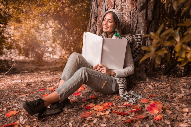 Mujer soñadora con libro sentado cerca de un árbol