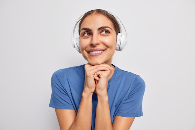 La mujer soñadora optimista mantiene las manos debajo de la barbilla mira hacia otro lado escucha música en auriculares estéreo y tiene pensamientos agradables viste una camiseta azul informal aislada sobre una pared blanca