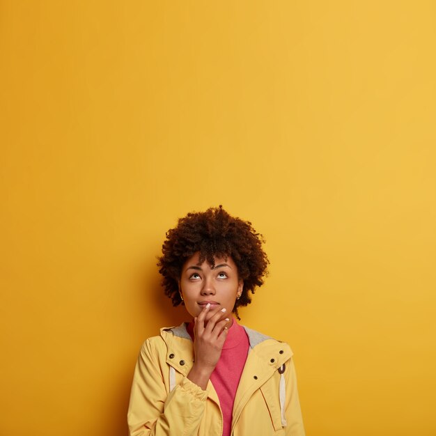 Mujer soñadora pensativa con cabello afro concentrado arriba, toma una decisión, mira pensativamente, mantiene la mano cerca de la boca, usa anorak amarillo