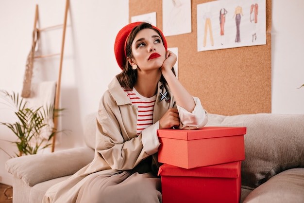 Mujer soñadora pensativa posando en la habitación y apoyado en caja de regalo. Señora pensativa en elegante boina roja y abrigo largo beige se sienta en el sofá.