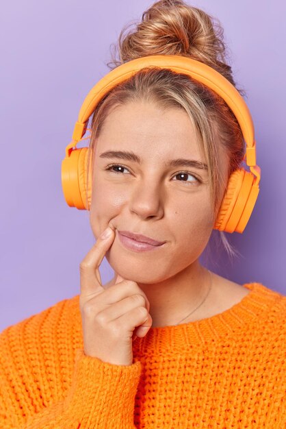 Mujer soñadora reflexiva mantiene el dedo índice cerca de la esquina de los labios sumido en pensamientos mientras escucha letras de canciones en auriculares vestidos casualmente aislados sobre fondo púrpura. Déjame pensar.