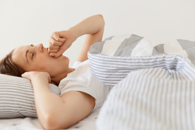 Mujer soñolienta con cabello oscuro con camiseta blanca casual acostada debajo de una manta y bostezando, tapándose la boca con el puño, simplemente despierta, disfrutando temprano en la mañana.