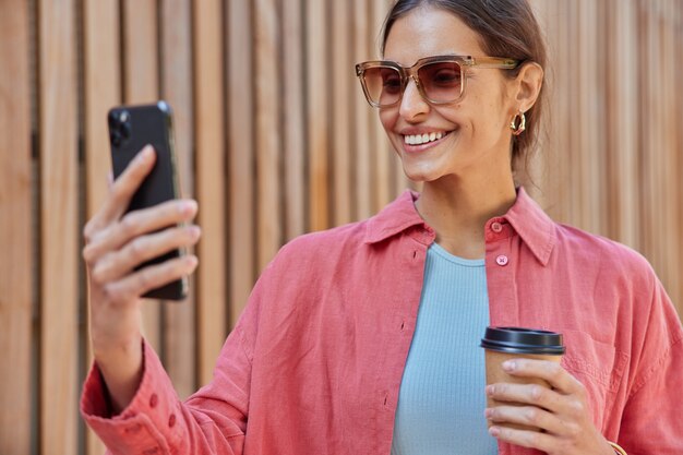 mujer sonríe ampliamente muestra dientes blancos usa gafas de sol camisa rosa sostiene el teléfono móvil frente a ella hace videollamadas bebe cafeína bebida de vaso de papel