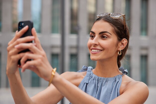 mujer sonríe con dientes graba video en el teléfono para su publicación en redes sociales hace llamadas en línea toma selfie usa ropa elegante posa al aire libre