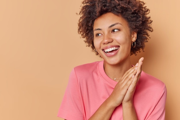 la mujer sonríe con los dientes mantiene las palmas juntas juntas mira hacia otro lado con alegría viste una camiseta casual aislada en el espacio de copia beige de distancia. Concepto de emociones positivas