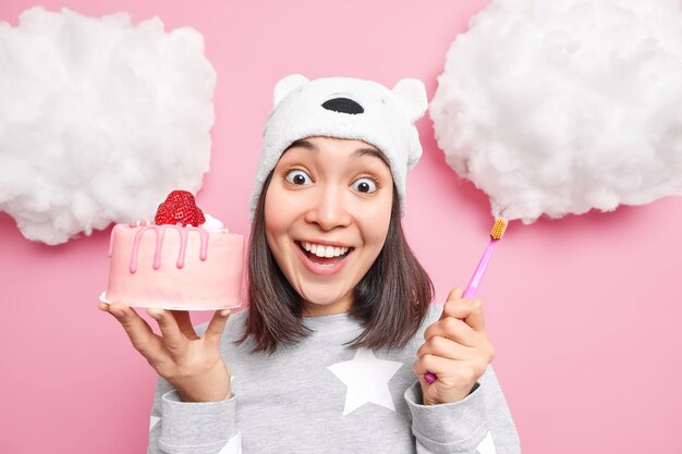 Mujer sonríe con dientes vestida con pijama sostiene un sabroso pastel y un cepillo de dientes tiene caries debido a comer demasiados dulces aislados en rosa