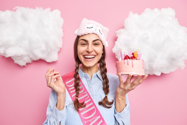 La mujer sonríe felizmente sostiene un delicioso pastel de fresa con velas celebra el cumpleaños disfruta de la fiesta doméstica usa una camisa casual con los ojos vendados en la frente aislada en la pared rosa