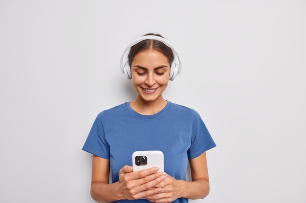 Foto gratuita la mujer sonríe positivamente sostiene el teléfono móvil descarga la canción a la lista de reproducción escucha la pista de audio en los auriculares viste una camiseta azul sobre blanco