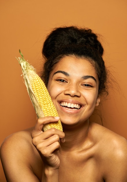 Foto gratuita mujer sonriendo a la cámara, sosteniendo mazorcas de maíz amarillo, posando en naranja