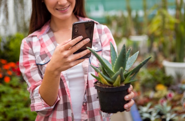 Mujer sonriendo tomando foto de planta con teléfono en invernadero