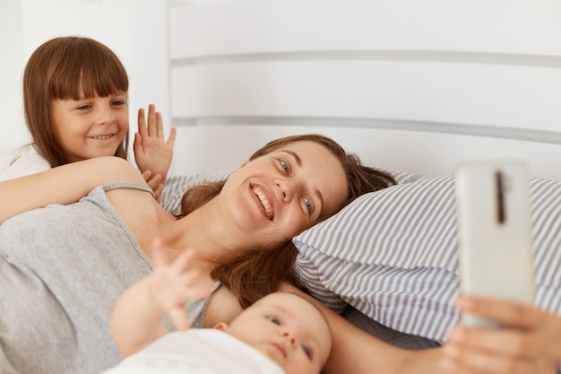 Mujer sonriente acostada en la cama con dos hijas, transmitiendo una transmisión en vivo temprano en la mañana o teniendo una videollamada, una niña mayor saludando con la mano a la cámara, saludando, diciendo hola.