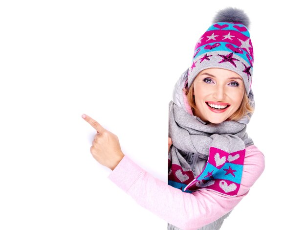 Mujer sonriente adulta en ropa de abrigo de invierno sostiene el cartel blanco en las manos y señala