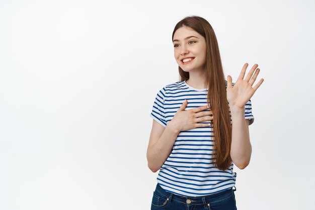 Mujer sonriente agitando su mano a un lado y luciendo amistosa, saludando a alguien, presentándose, parada sobre fondo blanco