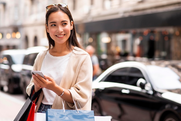 Mujer sonriente al aire libre con smartphone y llevando bolsas de la compra.