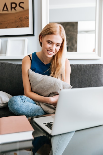 Mujer sonriente con almohada sentado y usando la computadora portátil en casa