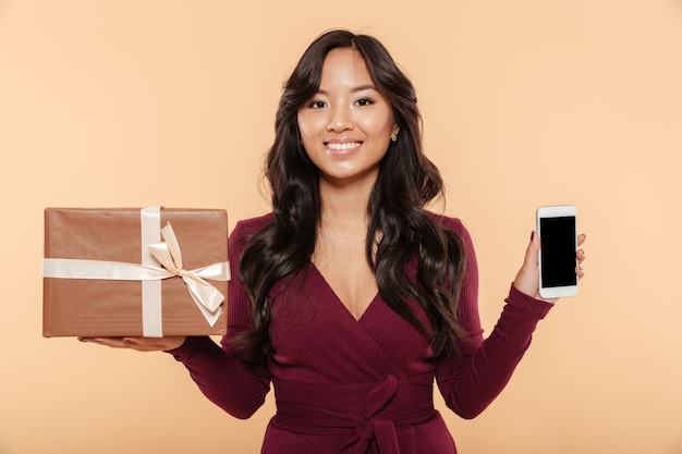 Mujer sonriente asiática en vestido marrón que muestra la caja actual con teléfono inteligente como regalo aislado sobre fondo de melocotón