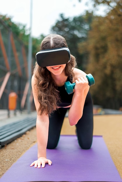 Foto gratuita mujer sonriente con auriculares vr en un chándal haciendo ejercicios con pesas en una estera de yoga en deportes