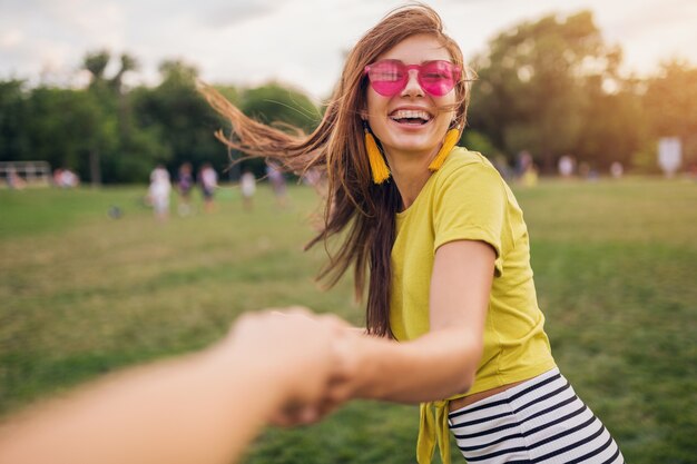 Mujer sonriente bastante elegante joven divirtiéndose en el parque de la ciudad, de la mano de su novio, sígueme, positivo, emocional, con top amarillo, gafas de sol rosas, tendencia de moda de estilo veraniego, emoción positiva