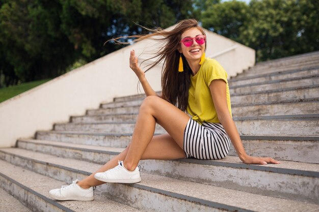 Mujer sonriente bastante elegante joven divirtiéndose en el parque de la ciudad, con top amarillo, minifalda, gafas de sol rosas, zapatillas blancas, tendencia de moda de estilo veraniego, piernas largas, sentada en las escaleras, agitando el pelo