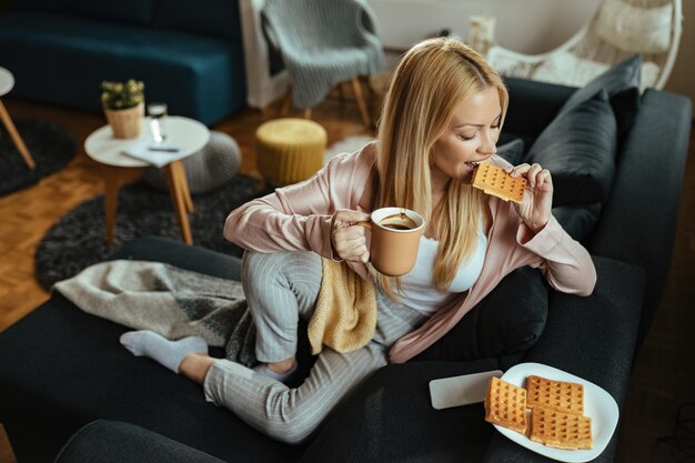 Mujer sonriente bebiendo café y comiendo gofres mientras se relaja en el sofá de casa