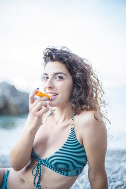 Mujer sonriente en bikini sentada en la playa aplicando bálsamo en los labios