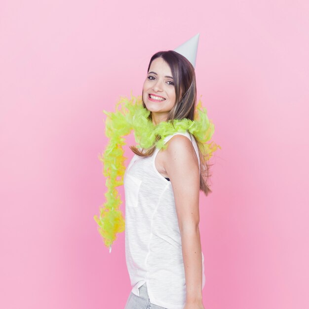 Foto gratuita mujer sonriente con boa de plumas verde alrededor de su cuello y sombrero de fiesta contra el fondo rosa