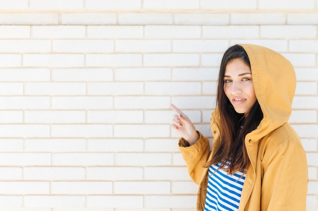 Mujer sonriente bonita que señala en la pared de ladrillo blanca