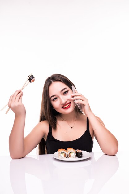 Mujer sonriente con cabello negro y labios rojos probar los rollos de suushi con palillos de madera en la mano hablando por teléfono