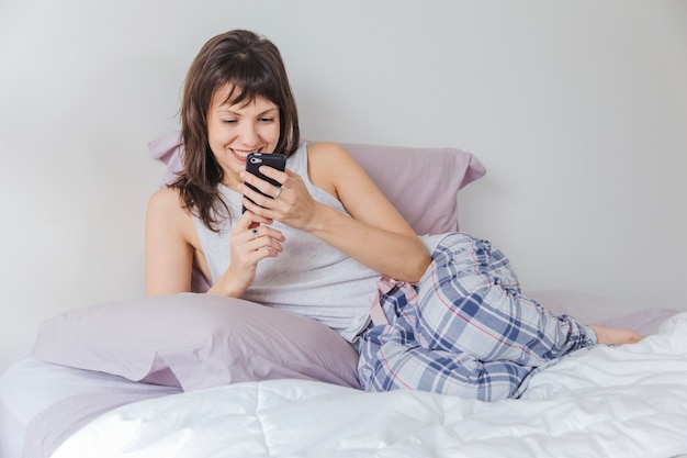 Foto gratuita mujer sonriente en la cama escribiendo en smartphone