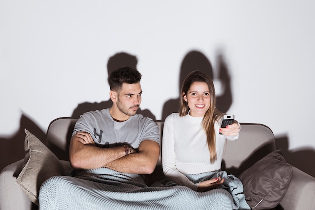 Mujer sonriente cambiando canales de TV cerca de hombre enojado en sofá