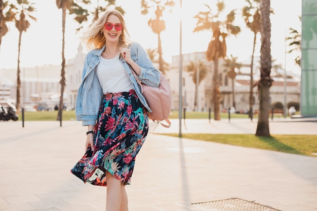 Mujer sonriente caminando en las calles de la ciudad con elegante falda estampada y chaqueta vaquera de gran tamaño con gafas de sol rosa