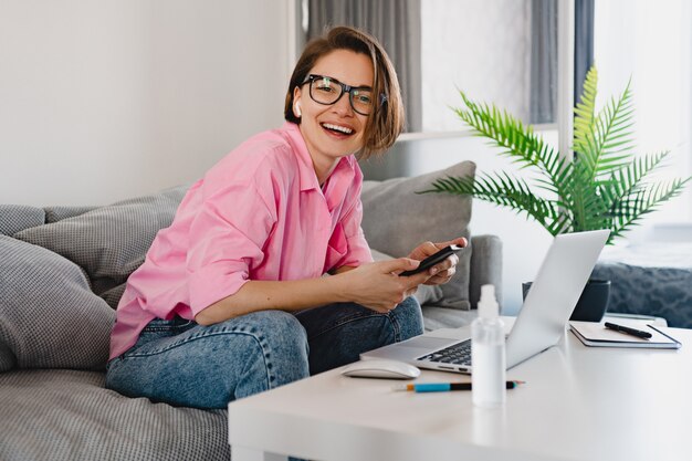 Mujer sonriente en camisa rosa sentado relajado en el sofá en casa en la mesa trabajando en línea en la computadora portátil desde casa