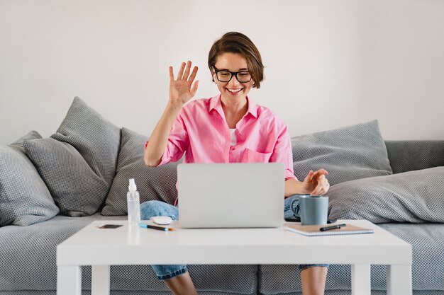 Mujer sonriente en camisa rosa sentado relajado en el sofá en casa en la mesa trabajando en línea en la computadora portátil desde casa