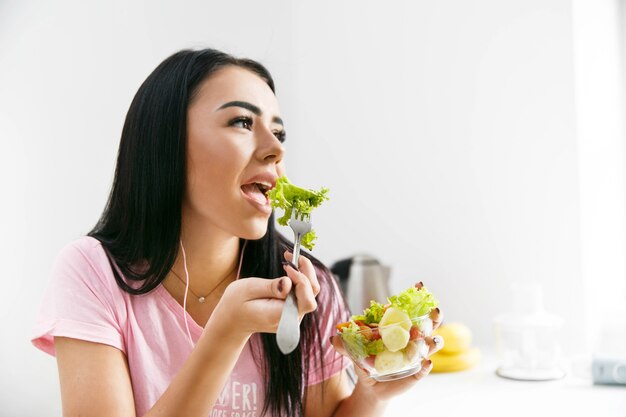 La mujer sonriente come la ensalada en la cocina blanca