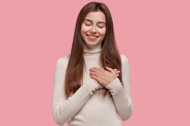 Mujer sonriente complacida mantiene las manos juntas sobre el pecho, expresa gratitud, mantiene los ojos cerrados