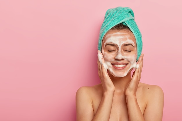 Foto gratuita mujer sonriente complacida que se lava la cara con gel limpiador, tiene jabón en la tez, mantiene los ojos cerrados, usa una toalla envuelta en la cabeza, tiene el cuerpo desnudo