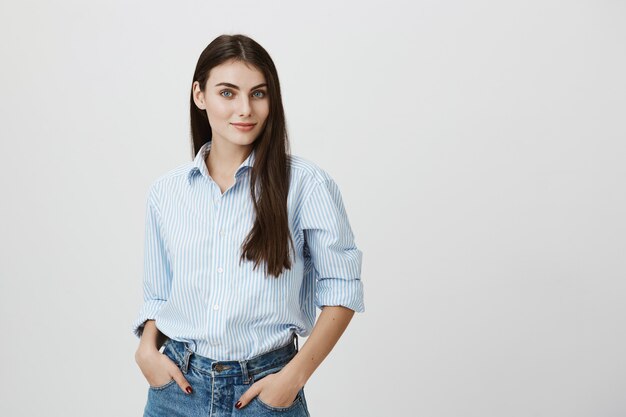 Mujer sonriente confiada en camisa y jeans, tomarse de las manos en los bolsillos