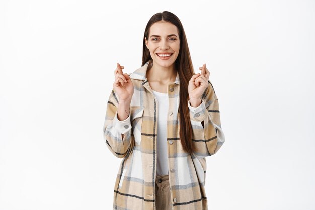 Mujer sonriente confiada cruza los dedos para tener buena suerte esperando noticias positivas ilusiones mirando a la cámara con cara determinada y anticipando estar de pie sobre fondo blanco