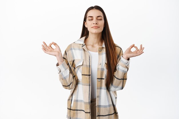 Mujer sonriente consciente meditando, respirando relajado con los ojos cerrados y pose de nirvana zen, practica yoga, descansando, sintiendo paz y alivio, de pie sobre la pared blanca del paciente